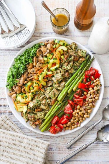 An overhead view of a grilled chicken salad salad that consists of rows of grilled pesto chicken, asparagus, mushrooms, and salads of cucumbers, tomatoes, and chickpeas.