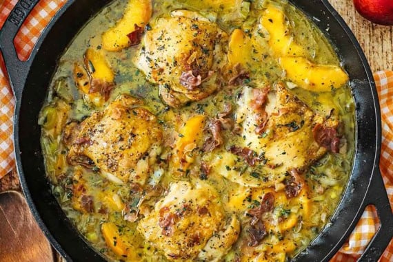 An overhead view of large black cast-iron skillet that is filled with braised chicken and peaches with a wooden spoon and fresh peaches nearby.