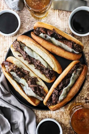 An overhead view of four large slow cooker French dip sandwiches with provolone cheese and several bowls of au jus sauce sitting nearby.