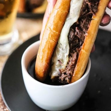 A close-up view of a slow cooker French dip sandwich being plunged into a small bowl of au jus sauce on a black plate.