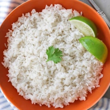 An overhead view of easy coconut rice that is in an orange serving bowl and has two lime wedges off to the side and a small sprig of cilantro resting on top.