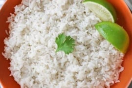 An overhead view of easy coconut rice that is in an orange serving bowl and has two lime wedges off to the side and a small sprig of cilantro resting on top.