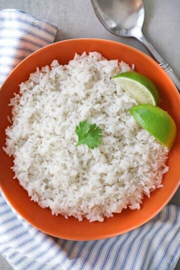 An overhead view of easy coconut rice that is in an orange serving bowl and has two lime wedges off to the side and a small sprig of cilantro resting on top.