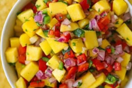 An overhead straight-down view of a small white bowl filled with fresh mango salsa with tortilla chips and limes wedges surrounding it.