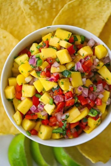 An overhead straight-down view of a small white bowl filled with fresh mango salsa with tortilla chips and limes wedges surrounding it.