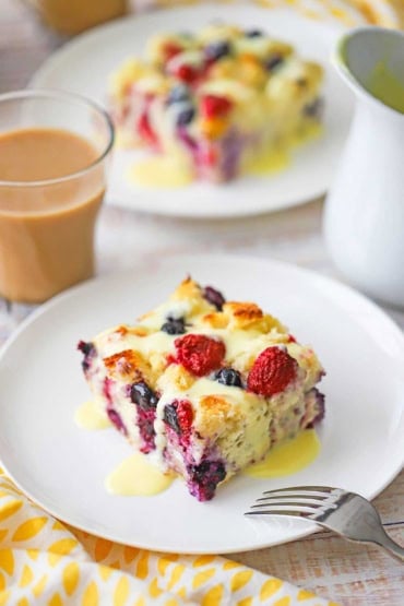 A straight-on view of a slice of mixed berry bread pudding with orange sauce sitting on a white dessert plate with a glass mug of creamed coffee in the background.