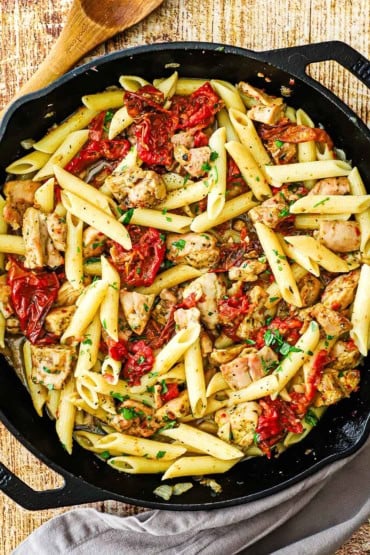 An overhead view of a large black cast-iron skillet that is filled with pasta with roasted tomatoes and pesto chicken.