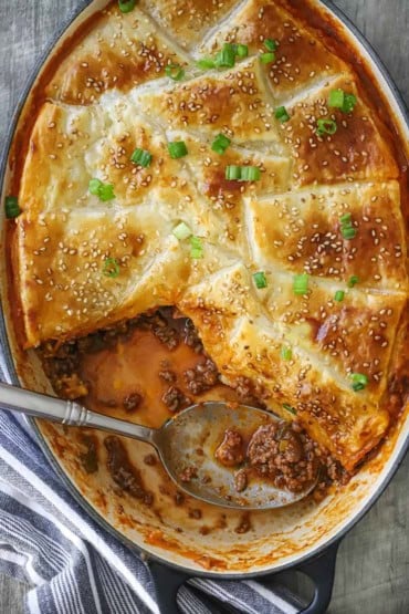 An overhead view of an oval baking dish filled with Sloppy Joe Casserole that is topped with a golden brown puff pastry and part of the casserole is gone and a spoon in the dish.