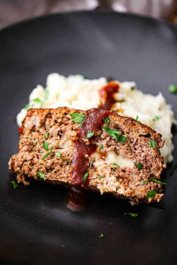 A close-up view of a slice of cheese-stuffed smoked meatloaf resting on a pile of mashed potatoes on a black plate.