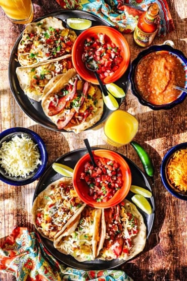 An overhead view of two large black plate filled with breakfast tacos that are stuffed with eggs, chorizo, bacon, potatoes, and sitting next to a bowl of pico de gallo.