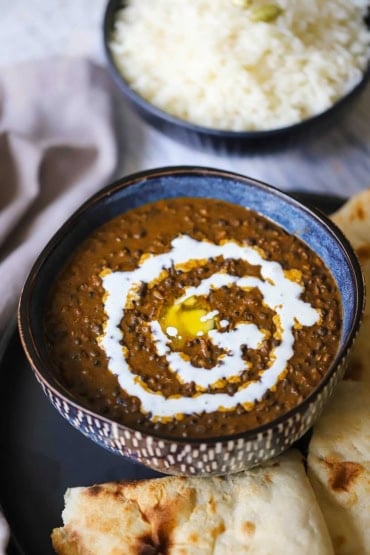 A straight-on view of a colorful blue bowl that is filled with a serving of dal makhani with another bowl of cooked basmati rice in the background.