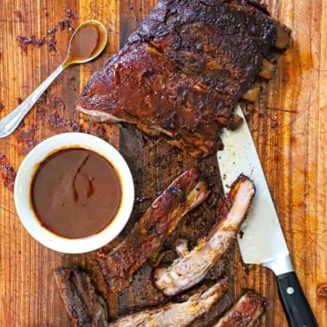 An overhead view of a rack of smoked St. Louis-style ribs that have been partially cut up with a bowl of homemade barbecue sauce nearby.