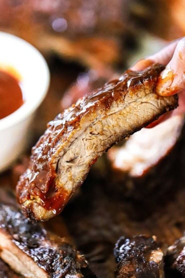 A person holding a single smoked St. Louis-style rib that has had homemade barbecue sauce brushed over the top of it.