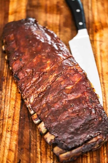 A straight-on view of a rack of smoked St. Louis-style ribs resting on a wooden cutting board with a large sharp knife next to it.