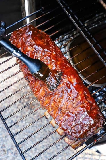 An overhead view of a rack of ribs that are resting on a rack in an electric smoker and are being brushed with BBQ sauce.