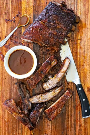 An overhead view of a rack of smoked St. Louis-style ribs that have been partially cut up with a bowl of homemade barbecue sauce nearby.