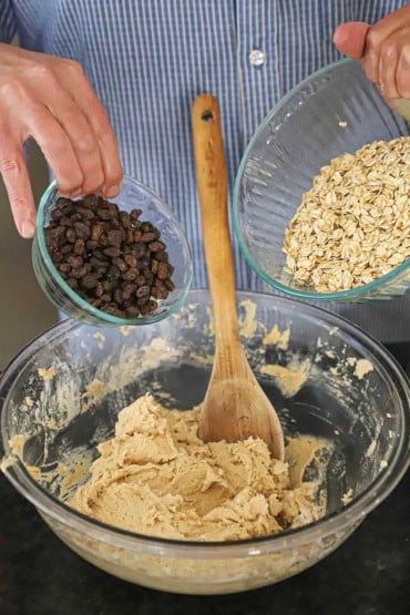 A person getting ready to dump a bowl of old-fashioned oats and another bowl of raisins into a third bowl filled with a cookie batter.