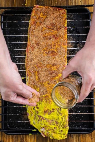 A person sprinkling pork rub over the top of a rack of uncooked St. Louis-style ribs that have a layer of yellow mustard smeared all over the ribs.