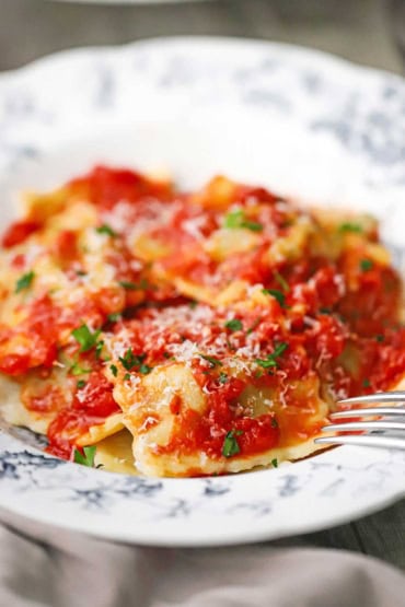 A close-up view of an antique bowl that is filled with a serving of homemade beef ravioli that is topped with a tomato sauce, shredded parmesan cheese, and chopped basil.