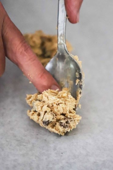 A person using his finger to push a tablespoon of oatmeal raisin cookie dough onto a baking sheet lined with parchment paper.