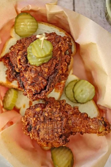 An overhead view of two pieces of Nashville hot chicken resting on slices of white bread with dill pickle slices scattered around all in a paper-lined basket.