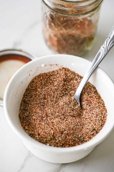 A close-up view of a small white bowl filled with best pork rub with a spoon inserted into the side of the rub.