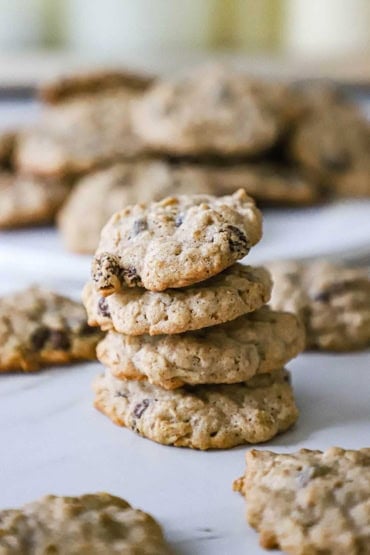 A straight-on view of four homemade oatmeal cookies stacked on top of each other on a piece of parchment paper with other similar cookies nearby.