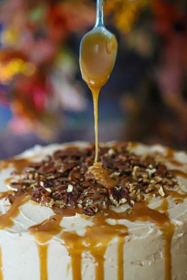 A spoon being used to drizzle homemade caramel sauce over the top of a caramel layer cake that is topped with chopped pecans.