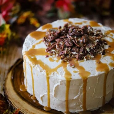 A straight-on view of a caramel layer cake that has homemade caramel drizzled over the tops and sides as well as circular pile of pecans on the top center of the cake.