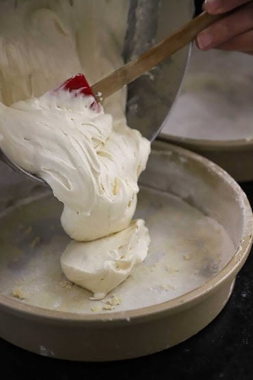 A person using a rubber spatula to transfer white cake batter from a mixing bowl into a 9-inch cake ban that has been greased and floured.