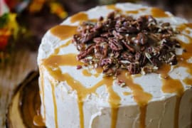 A straight-on view of a caramel layer cake that has homemade caramel drizzled over the tops and sides as well as circular pile of pecans on the top center of the cake.