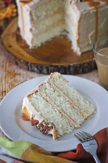 A straight-on view of a single serving of caramel layer cake on a white dessert plate with the cake in the background.