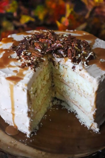 A straight-on view of a caramel layer cake sitting on a wooden cake platter and the cake has a large portion of the cake missing revealing the layers of white cake and caramel frosting.