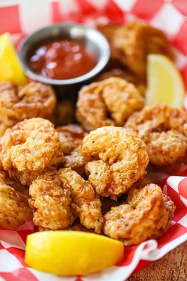 A close-up view of Southern-fried shrimp in a basket that is lined with red checkered paper and a bowl of cocktail sauce in the background.