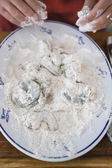 A person tossing seasoned shrimp in all-purpose flour on a large oval platter.