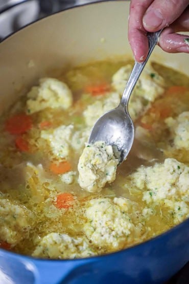 A person using a silver spoon to transfer uncooked dumplings dough into a Dutch oven filled with chicken broth, carrots, and other mounds of dough.