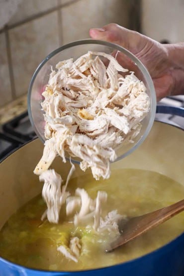 A person dumping shredded roasted chicken from a glass bowl into a large Dutch oven that is filled with a simmering chicken broth.