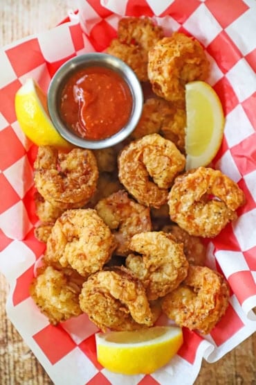 An overhead view of a basket lined with a red checkered paper and filled with Southern fried shrimp with a container of cocktail sauce and lemon wedges nearby.