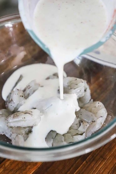 A person pouring a buttermilk marinade over uncooked shrimp in a large glass bowl.