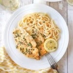 An overhead view of two cutlets of chicken piccata topped with lemon and capers sauce and flanked by a serving of cooked linguine and a slice of lemon.