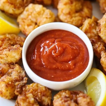 A close-up view of a small white bowl filled with homemade cocktail sauce surrounded by fried shrimp and lemon wedges.