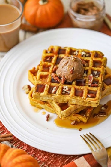 A straight-on view of a stack of three pumpkin waffles that are topped with a dollop of whipped cinnamon honey butter, maple syrup, and chopped pecans.