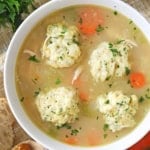 An overhead view of a white soup bowl that is filled with homemade chicken and dumplings including sautéed carrots and celery.