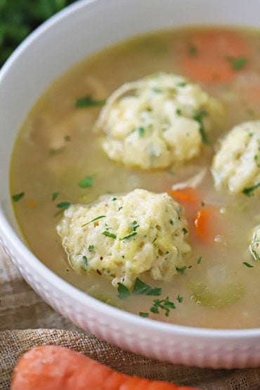 A close-up view of a white soup bowl that is filled with homemade chicken and dumplings soup that has been garnished with chopped fresh parsley.