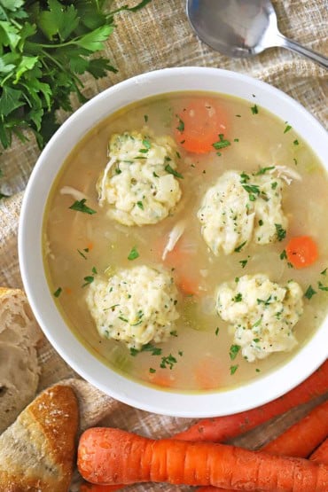 An overhead view of a white soup bowl that is filled with homemade chicken and dumplings including sautéed carrots and celery.