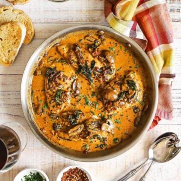 An overhead view of a large silver skillet filled with creamy Tuscan chicken with slices of toasted bread and a bowl of grated cheese nearby.