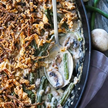 An overhead view of a baking dish that partially filled with gourmet green bean casserole with a serving spoon resting in the dish next to saucy green beans.