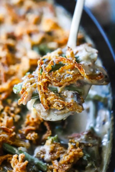 A close-up view of a large serving spoon holding up a helping of homemade green bean casserole with fried onion strips on top.