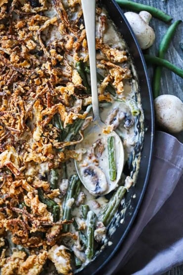 An overhead view of a baking dish that partially filled with gourmet green bean casserole with a serving spoon resting in the dish next to saucy green beans.