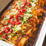 A close-up view of leftover turkey enchiladas in a white baking dish with a red sauce and topped with chopped lettuce and pico de gallo.